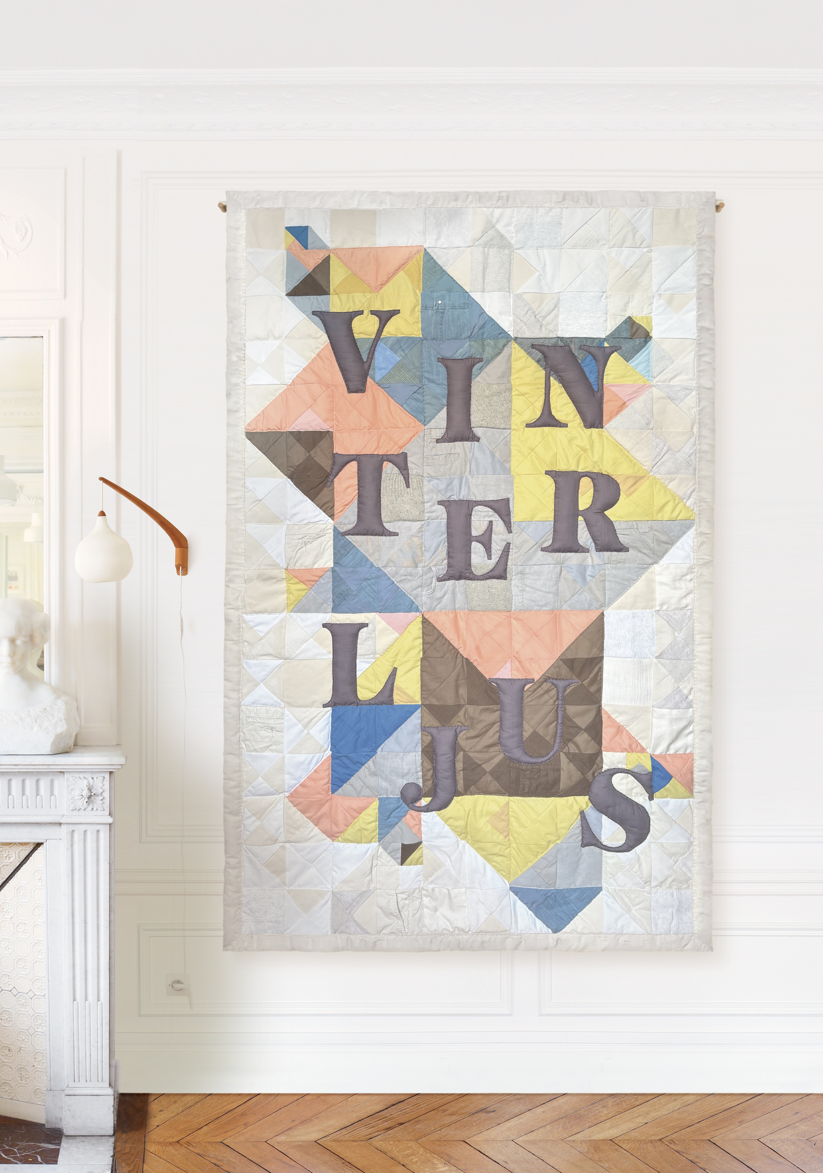 Textile artwork in shades of grey, blue, taupe, pink, pale yellow and off-white with hanging grey letters in an apartment – memory patchwork by Pénélope Westberg